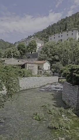 A river flows past stone walls and buildings on a hillside. Trees and vegetation surround the buildings. The sky is partly cloudy.