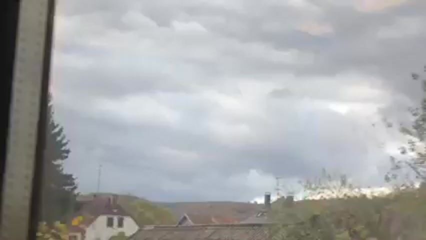 The video appears to be taken from inside a moving train or vehicle, looking out the window. The scenery includes houses with brown tiled roofs, trees, and a cloudy, overcast sky. The camera movement suggests the train is in motion.