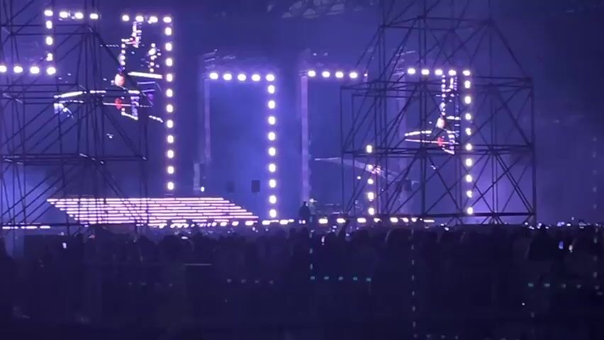 The video shows a large indoor concert venue with a massive stage setup. The stage features a complex structure of metal scaffolding supporting large, illuminated displays forming geometric shapes and what appear to be numbers. These lights are primarily purple and white. There are multiple screens visible, some displaying abstract patterns and others showing close-ups of performers. A DJ booth is visible on a raised platform on the left side of the stage, and a DJ is performing. Smoke or fog is present on stage, creating atmospheric effects. In front of the stage, a large crowd of people is gathered, silhouetted against the stage lights, with many holding up their phones to record. The overall lighting is dark with vibrant stage lighting dominating the scene.