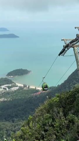 The video pans from a lush green, forested mountainside, offering a high-angle view of the surrounding landscape. In the foreground, the edge of the mountain is visible, covered in dense foliage. A bright green cable car is the main focus, suspended by a thick cable that stretches across the frame. The cable car is moving away from the viewer, traversing the expanse between the mountain and a distant body of water.

Below the cable car, a coastline unfolds, characterized by a strip of sandy beach transitioning into a developed area with buildings. Further out, the turquoise water stretches to the horizon, dotted with several small, verdant islands. In the distance, more islands and a hazy coastline are visible under a sky with scattered clouds. The overall impression is one of a scenic aerial view, capturing a cable car journey over a tropical island landscape with the sea and other islands in the background.