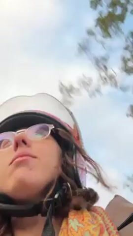 A person wearing a helmet and glasses is riding a motorbike, looking at the camera and talking. Behind them are green trees and hills, with a cloudy sky above. The person is wearing a patterned scarf and brown jacket. The wind is blowing.