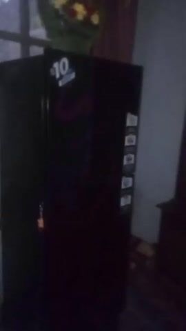 The video shows a dark red refrigerator with a "10" sticker on top, standing in a dimly lit room. The room appears to be a bedroom or living area, with a dark wood dresser visible to the right of the refrigerator. A floral-patterned rug is on the floor. The camera pans slightly to the left, showing a wall with an electrical outlet and some peeling paint. There is a window in the background.