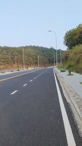 The video shows a newly paved road winding uphill. The road has a white line marking the edge and dashed white lines dividing the lanes. There is a solid yellow line near the center. Streetlights are spaced along the left side of the road, with a hillside covered in trees beyond them. The sky is clear and light blue. A concrete curb and grassy area are visible on the right side of the road. A person is visible in the distance, standing by the side of the road.