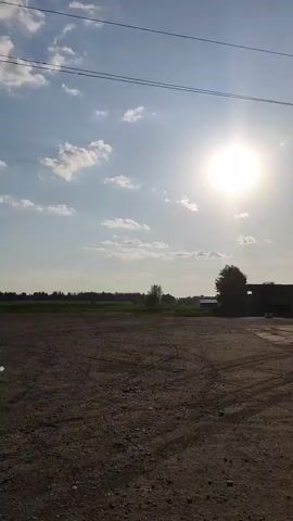 The video presents a static, medium shot of a rural landscape under a bright, sunny sky dotted with scattered clouds. The foreground is dominated by a gravel-covered ground, displaying numerous tire tracks. In the middle ground, there is a grassy field and a line of trees, forming a natural boundary. A road with signage is visible on the left side of the field. Above, power lines stretch across the top of the frame. The sun is positioned high in the sky, slightly to the right, emitting a strong glare.
