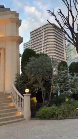 The video shows the exterior of a building with the word "ANNIVERSAIRE" above the entrance. The building is light-colored with a classical design, featuring columns and a staircase leading to the entrance. There's a stained-glass window visible near the door. The surrounding area is decorated with trees, bushes, and ornate light fixtures. Above the walkway are strings of globe-shaped lights. The video also captures a building with a spire in the background and flags bearing the "ANNIVERSAIRE" logo. The sky is visible in the background.