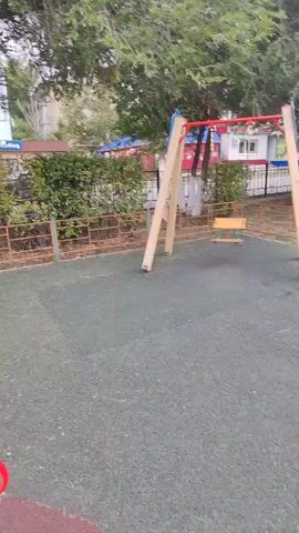 The video pans across a children's playground. In the foreground, a red merry-go-round with a yellow seat is visible. A young girl holding an ice cream cone stands next to it. Further back, a woman in dark clothing sits on a brown bench shaped like a bear. Behind her, a multi-colored climbing structure with a pink frame stands on a red and yellow rubber surface. Beyond the playground, there are flower beds with colorful flowers, trees, a seesaw, and a building in the background. The sky is overcast.