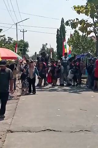 This is a short video of an outdoor street scene, possibly a parade or festival. The pavement is cracked and uneven. On the left side of the road, a person in a dark shirt walks away from the camera. Further down the road, there are tents and a crowd of people. Several individuals are dressed in large, bulky costumes that appear to be robotic or armored. Some of these costumes are dark gray and have intricate designs. There are also people in casual clothing, including women and children. Many people are looking towards the costumed figures. Flags, some red and white, and others with green, yellow, and red stripes, are visible in the background, attached to poles or held by individuals. The overall atmosphere seems lively, with a gathering of spectators. The lighting suggests it is daytime.