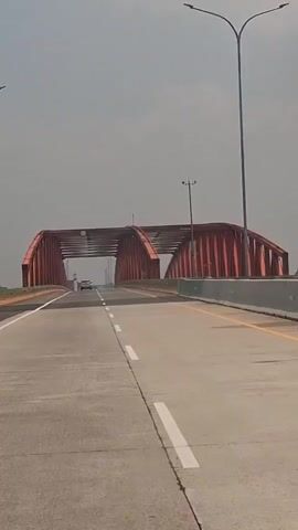 A vehicle approaches a large red bridge with an arched structure over a highway. The sky is overcast, and the road appears to be made of asphalt with white lane markings. There is one other vehicle visible on the bridge, moving away from the camera.