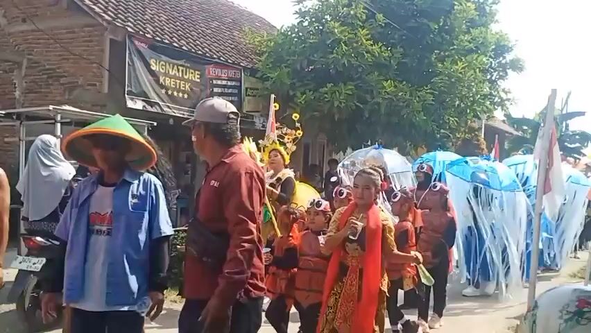 A group of children in matching costumes, some dressed as jellyfish with blue umbrella-like headpieces and white streamers, others in brown and orange costumes with large circular eye accessories on their heads, participate in a parade. A woman in a yellow costume with a sunflower headdress walks alongside them. Another person in a maroon shirt and a grey baseball cap is visible in the foreground, facing away from the camera. A building with a banner reading "SIGNATURE KRETEK" is present on the left side of the frame. The parade appears to be taking place on a street with houses and trees in the background. The sky is bright and sunny.