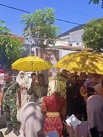 A group of people are gathered outdoors under a clear blue sky. Two large, ornate yellow umbrellas are held above a central figure. The central figure appears to be a woman adorned in a richly decorated red and gold garment and a distinctive headpiece. She is seated and surrounded by people. To the left of the central figure, a man in military camouflage uniform stands facing the crowd. Several other individuals are visible, some wearing headscarves and traditional clothing. In the background, a white building with a dark roof and trees are present. The scene appears to be a procession or a ceremonial event.