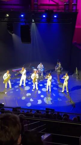 A live musical performance is captured from a high angle, looking down onto a stage. The stage is lit with blue and purple lights, with a spotlight casting a bright beam from above. A band of seven musicians is performing on stage. Five musicians in the front row are wearing matching white uniforms and playing brass instruments, appearing to be saxophones and possibly a trumpet or trombone. A drummer is seated at the back of the stage behind a drum kit. To the right of the drummer, a musician in a dark uniform with red trim, possibly a military uniform, is playing bagpipes. The stage floor has a circular pattern of light reflected on it. In the foreground, the audience is visible, with their backs to the camera, seated in rows of dark seats. The overall environment appears to be an indoor venue, likely a concert hall or theater.
