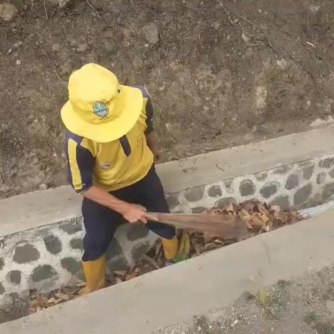 A person wearing a yellow hat and a yellow and blue shirt is sweeping leaves out of a concrete ditch with a broom. The ditch has stone walls. The person is also wearing yellow boots and dark pants. The ground above the ditch is dirt and rocks.