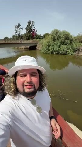 A man takes a selfie video on a wooden bridge. He is wearing a white t-shirt, a white Nike bucket hat, and sunglasses hanging from his shirt. He has curly brown hair and a beard. A river runs alongside the bridge. The water is a muddy brown color. Bushes and trees grow along the river bank. In the background, another bridge can be seen. The sky is bright and clear. The man says, "This is me. This is the river."