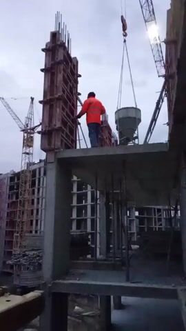 The video shows a construction site under a cloudy sky. A construction worker in an orange jacket and dark pants stands on a concrete platform, using a long metal tool to adjust a tall, rectangular concrete formwork. A crane is positioned above the platform, holding a large, cone-shaped container, presumably for pouring concrete. Another crane is visible in the background, along with the partially constructed frame of a multi-story building. The concrete platform is supported by vertical metal beams.