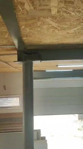 The video shows a close-up of a bird's nest built on a metal beam and supporting column. The nest is made of mud and straw. A bird, possibly a swallow, is perched on the edge of the nest. The background shows a wooden ceiling and a large overhead door, suggesting the location might be a garage or industrial space. The lighting is natural, coming from an unseen source. The camera is steady, focusing on the nest and bird.