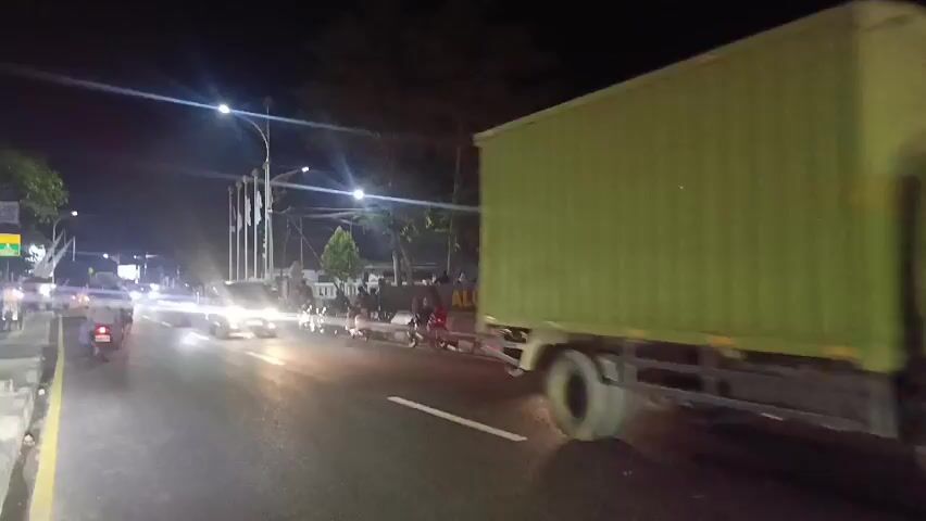 The video shows a nighttime street scene with traffic. In the foreground, a person on a motorcycle is riding down the road. A large truck is also visible, as is a bus with decorative lights. The street is lit by streetlights and the headlights of vehicles. There are some people standing on the side of the road, and a sign that reads "ALUN ALUN".