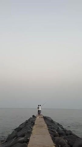 Two people stand at the end of a stone jetty extending into the sea, with one individual holding a fishing rod and casting a line into the water. The sky is overcast, creating a soft, diffused light over the scene. The sea is calm with small ripples, and the jetty is constructed of large dark rocks topped with a paved pathway.