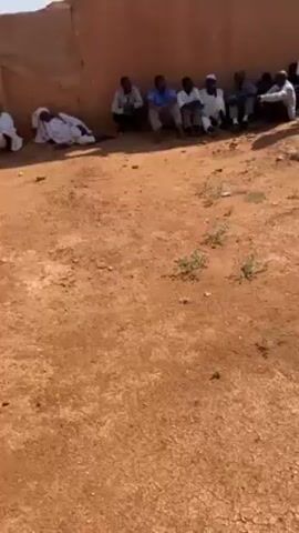 The video shows a group of people, mostly men, sitting against a wall in a dirt area under a sunny sky. They are likely Sudanese, given the traditional attire and environment. There's a ruined building in the background. A woman in black walking across the space in front of the group. A minaret is visible in the background. The atmosphere appears calm and static.