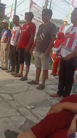 This is a shaky, low-angle, outdoor video that appears to be filmed in daylight. Several people, mostly men, are standing in a line, facing away from the camera, with their heads tilted back as if looking up. They are dressed in casual clothing, including t-shirts, shorts, and sandals. Some of the men are wearing shirts with the number "81" and the word "INDONESIA" on them, suggesting they may be part of a team or group. There are flags and banners in the background, including Indonesian flags. The ground is paved with concrete. In the foreground, someone's legs are visible, lying down, and they are wearing red pants. Another person in the background is wearing a red hijab and red clothing. The overall atmosphere seems to be that of an outdoor event or gathering.