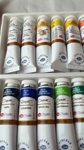 The video is a close-up, top-down shot of a set of oil paint tubes arranged in a white plastic tray. The tubes are neatly organized in two rows, with the top row filled with colors like yellow, orange, and red, and the bottom row featuring blues, greens, and browns. Each tube has a white cap and a label with brand information, color name, and a barcode. The lighting is bright and even, highlighting the colors and details of the paint tubes. The background is a neutral, light-colored surface, possibly a table or a cloth. The camera slowly pans across the tray, allowing the viewer to see the entire collection of paints. There are no people or any other objects visible in the frame.