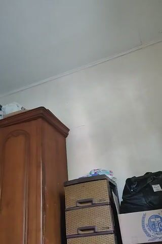 Here is a forensic description of the video:

The video presents a low-angle, static shot of the interior of a room. The focal point is on a corner where the wall meets the ceiling.

**Objects:**
- A wooden dresser, brown in color, is positioned in the lower portion of the frame.
- Stacked on top of the dresser are items including a small cabinet with woven textures and brown handles, a box with the word "DEWA" printed on it, and a black bag.
- The ceiling has a visible window frame.
- A green piece of fabric is draped across the window frame, obscuring the view outside.
- A dark round object, possibly a light fixture, is attached to the ceiling near the window.
- The walls are white or a light color.

**Environment:**
- The room appears to be somewhat cluttered with items stored on top of the dresser.
- The lighting is ambient, with some natural light filtering through the window.

**Overall Impression:**
The scene seems to be a storage space or room with limited natural light and a mix of furniture and stored items.