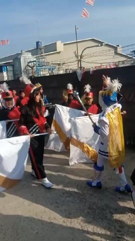 A short video shows a small marching band moving along a paved area beside a wall, with industrial buildings visible in the background.  The band consists of approximately 10-12 young people wearing red and black uniforms with feathered hats. Some are carrying and waving white flags with gold trim, while others are playing drums and cymbals.  In front of the band is an individual dressed in an elaborate white and blue costume with a gold cape, carrying a baton. One man walks next to the band, wearing a black t-shirt, sunglasses, and gray pants.  Above the buildings, a few Indonesian flags are suspended.  The sky is blue.
