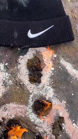 A black beanie with a white Nike logo rests on a rock. Several small piles of dark material are on the rock, some of which are on fire. There is white powder sprinkled around the material. A green coconut is placed in front of the burning material. Two large curved horns are visible on either side of the coconut. Black fabric, possibly clothing, is underneath the coconut and horns.