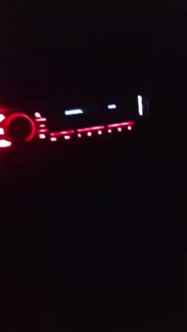 The media file captures a close-up view of a car stereo system in a dark environment. The stereo's display screen shows text, though it's partially obscured and difficult to read definitively. The illuminated elements of the stereo are primarily red and white, suggesting it is powered on. The surrounding darkness indicates it is likely nighttime. The scene is static, focusing solely on the car stereo. There are no discernible people or broader environmental details visible, other than the implied interior of a vehicle due to the presence of a car stereo. The text on the screen appears to be a combination of letters and numbers, possibly indicating a radio station or track information. The overall impression is one of a dimly lit interior with the car stereo as the sole focal point.