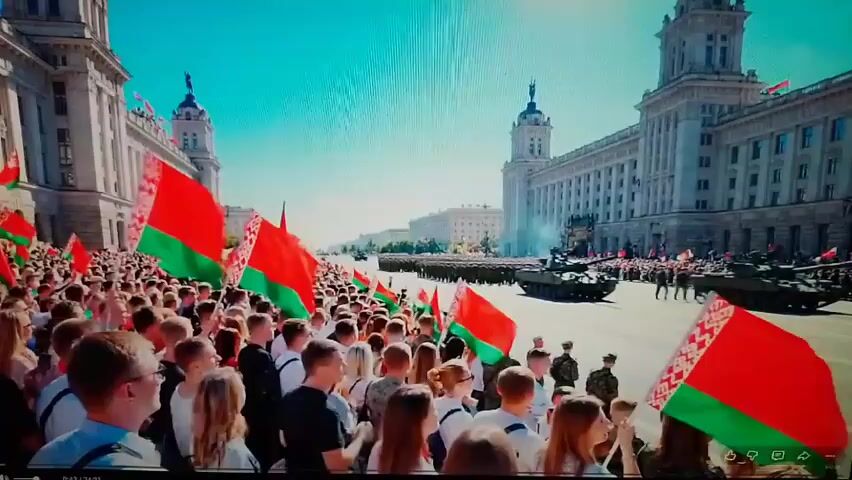 This video depicts a parade with a large crowd of people on a street, holding many red and green flags. Military vehicles, specifically tanks, are visible in the background, along with soldiers marching. The buildings on either side of the street are large and ornate, suggesting an official or ceremonial event. The sky is clear and blue, indicating it is daytime. The crowd appears to be cheering and waving their flags, suggesting an enthusiastic reception of the parade.