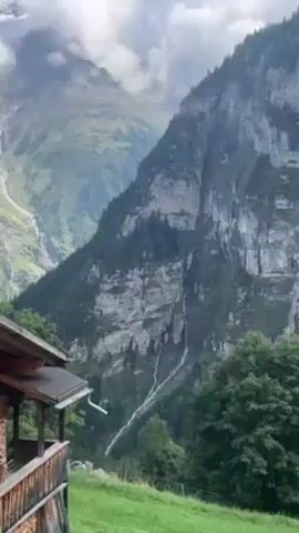The scene captures a mountainous landscape, likely in the Swiss Alps, under a partly cloudy sky. Dominating the background are towering mountain cliffs with textured rock faces, interspersed with patches of green vegetation and evergreen trees. A slender waterfall cascades down the side of one cliff. In the foreground, the corner of a traditional wooden chalet is visible with a brown roof and timbered walls. A green lawn extends in front of the chalet, bordered by lush trees. The overall impression is one of serene beauty in a mountainous setting.