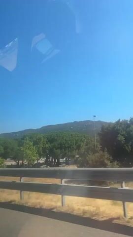 The video is a first-person perspective, likely from a vehicle, looking out a window. The sky is a clear, bright blue. In the foreground, there is a metal guardrail. Beyond the guardrail, there is dry, yellow grass. Further in the distance, there are trees, some appearing to be pine trees with conical shapes. Behind the trees, a gently sloping mountain range is visible, sparsely covered with vegetation and dotted with what appear to be numerous light poles. To the right, a white building with a sign that says "COMO" in large black letters is visible. The Burger King logo is also prominently displayed on this building. There are other smaller structures and some vehicles visible in the vicinity of the building. The overall scene depicts a roadside view with a clear sky, natural landscape, and commercial establishments.