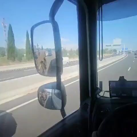 From the interior of a truck, the driver views a highway. The driver is passing military vehicles on the highway. The driver-side mirror shows more military vehicles behind the truck. The weather is sunny. The landscape to the left shows some trees.