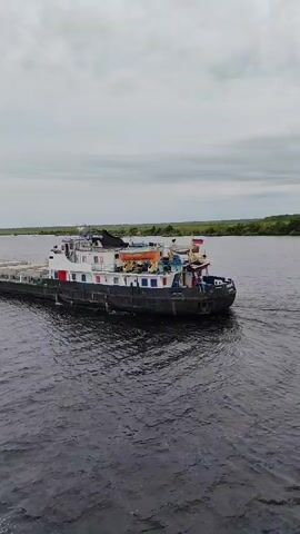 The video shows a long, dark barge with a white superstructure, carrying cargo, moving slowly on a wide, calm river under an overcast sky. Green trees line the distant bank of the river. The camera is positioned at a slightly elevated angle, possibly from another boat, capturing the barge from its stern quarter. A small amount of white spray is visible at the stern of the barge, indicating movement. The barge appears to be a cargo vessel. There are no visible individuals on the deck of the barge, although there are some colorful objects scattered across the deck, suggesting the presence of crew or equipment. The overall scene is serene and depicts a typical day of river transportation.