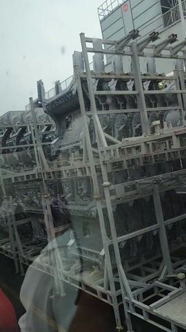 The video shows a close-up view of car parts, likely chassis components, stacked on metal racks. The parts are silver and appear to be made of metal. They are arranged in a multi-layered structure, suggesting they are being transported or stored. The camera is tilted, giving an upside-down perspective of the objects. Reflections on a surface, likely a window, show a person's head and shoulders, with a white garment and a red object in the foreground. The background is overcast and appears to be an outdoor industrial or storage area. The scene is filmed from a moving vehicle, as indicated by the slight blur and the changing reflections. There is some Chinese text visible on the metal racks, though it is not clearly legible. The weather appears to be gloomy. The overall impression is one of industrial activity and logistics, with the focus on the manufactured car parts.