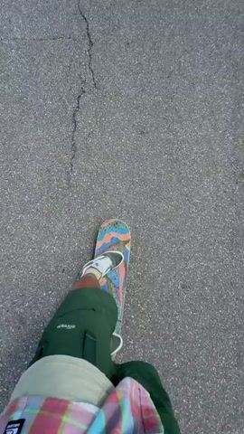A person wearing green pants and white sneakers is skateboarding on a paved road. The road is cracked in several places. The skateboard has a colorful design. The scenery changes to a wide-angle shot of a winding road through a green, grassy field with hills in the distance under a blue sky with some clouds. The camera then returns to a close-up of the skateboard and the road.