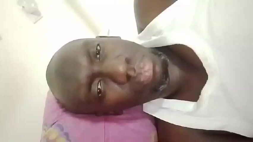 A close-up shot features a dark-skinned, bald man lying on his left side on a pink pillow. He is wearing a white shirt. He is holding his right hand up and pointing his index finger toward the camera with his lips pursed as if to make a "shush" gesture. A white wall is visible in the background, with a white electrical outlet cover in the upper left corner.