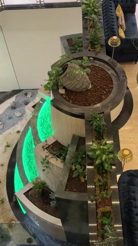 An aerial shot shows a tiered indoor water feature with lush green plants. The water feature is made of dark stone and has a curved design, with illuminated green lights along the lower tiers. A large, ornate ceramic pot is placed at the top of the water feature, spilling green vines over its edge. The surrounding area appears to be a modern lobby or lounge, with dark tufted sofas and decorative lamps visible in the background. The floor is a light-colored tile, and a pathway of smooth stones leads away from the water feature.