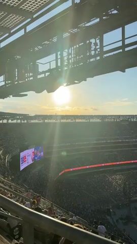 An overhead shot looks down on a packed stadium, the audience spread across green seats. In the distance, a large pink screen displays "REQUEST A SONG" and a cassette tape graphic, flanked by QR codes. Above the screen, a digital scoreboard shows a game in progress with players in blue uniforms and text that reads "MOODY'S" and "NYSK." The sun, a bright white orb, shines in the upper left quadrant of the sky, casting a lens flare that streaks across the image. The stadium structure, a dark metallic framework, hangs over the top of the frame.