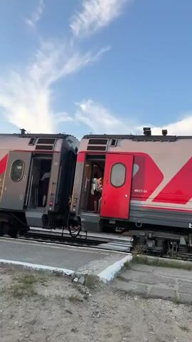 A woman in a uniform stands at the open doorway of a train car. The train appears to be stopped at a station, as evidenced by the platform visible in the foreground. The sky above is blue with white clouds, suggesting it is daytime. The woman is looking out of the doorway and appears to be interacting with someone inside the train or on the platform. The train cars are gray and red, with Russian text visible on the side.