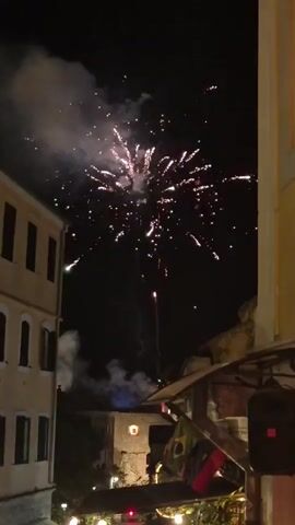 The video shows a nighttime scene with fireworks exploding in the sky. The fireworks are colorful, with flashes of white, green, and red. Smoke from the explosions drifts across the sky. In the foreground, there are buildings with windows. Some of the buildings appear to be old, with traditional architecture. There are also some decorations visible, including string lights and flags. The overall impression is of a celebration or festival.