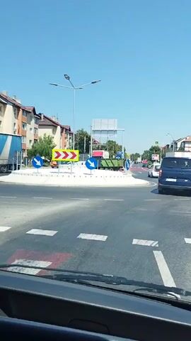 The video shows a view from the driver's seat of a vehicle approaching and navigating a roundabout on a sunny day. The roundabout has several directional signs, including mandatory turn arrows. Traffic signs for a roundabout are visible, with blue circular signs indicating the direction of travel. A number of vehicles are present in the roundabout and on the roads leading to it, including cars, a van, and a truck. Buildings line the streets, and trees are visible in the background. The road surface is asphalt with white lane markings and pedestrian crossings. The sky is clear and blue. The camera is positioned to capture the road ahead, the roundabout, and the surrounding environment.