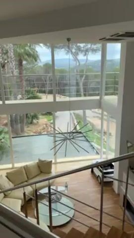 The video shows the interior of a modern-style house, likely a living room, taken from an elevated position such as the top of stairs. The room features a large window wall that stretches two stories high, providing an expansive view of the outside. The exterior features palm trees, other green foliage, and rolling hills in the distance. In the foreground, a cream-colored sectional sofa, a glass-topped round coffee table, and a complex, spiky, black chandelier are visible. The floor is made of wooden planks. A small white toy car sits near the staircase. The camera slowly pans upwards, focusing more on the large window.