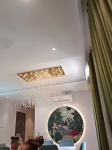 The camera is positioned low, looking up at a well-lit room with a high white ceiling. A large, rectangular chandelier hangs from the ceiling, featuring a golden base and numerous clear, teardrop-shaped crystals suspended below. Several recessed lights are also visible in the ceiling. A large, round piece of art depicting a floral scene with greenery and flowers adorns the wall. To the right, there is a floor-to-ceiling curtain in a light green color. At the bottom of the frame, the tops of several heads are visible, suggesting people are sitting or standing just out of view. An air conditioning unit is mounted on the wall.