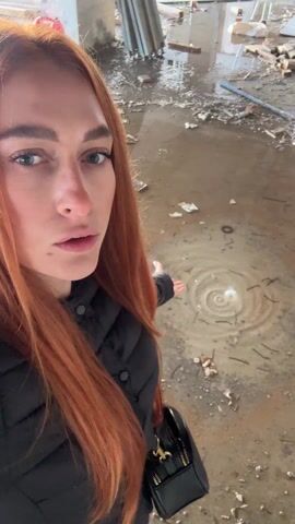 A person with long red hair wearing a black quilted jacket and carrying a small black purse stands in an unfinished or dilapidated building with a water-covered floor. The floor is littered with debris. The person gestures towards a spot in the water where circular ripples are visible, seemingly caused by water dripping from above. The person looks directly at the camera with a surprised or concerned expression.