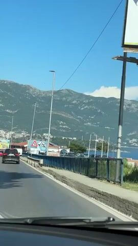 This is a first-person perspective video of a car driving over a bridge on a sunny day. The scene is a coastal road with a view of a bay, mountains, and a clear blue sky. The time of day appears to be midday.

In the foreground, the dashboard and windshield wipers of the car are visible. The road is asphalt, and a concrete barrier with a blue metal fence runs along the right side of the bridge. Beyond the fence, there's a sidewalk, some greenery, and a view of the water. Several poles, likely for streetlights, are spaced along the bridge.

In the mid-ground, across the bay, there is a town nestled at the foot of a large, rugged mountain range. The mountains are covered in greenery, and some buildings are visible on their slopes. A few boats are scattered across the water.

In the background, the imposing mountains dominate the landscape. The weather appears to be clear and warm, typical of a summer day. There is a red and white triangular sign with a black symbol on it visible on the left side of the road, indicating a potential hazard. A white van is also visible on the road ahead. The overall mood is one of travel and scenic enjoyment.