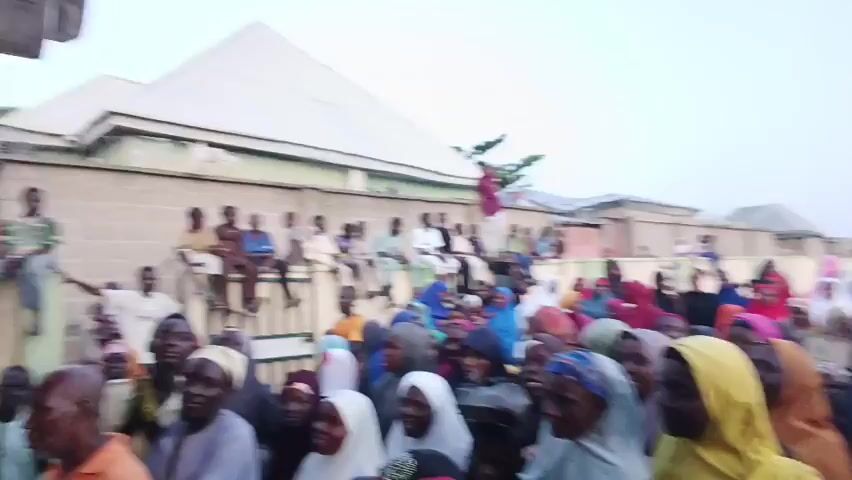 A crowd of people is gathered in what appears to be a village or a densely populated area. Many women in the foreground are wearing hijabs in various colors, including white, blue, yellow, and red. Some men are visible among them. Children are sitting on a wall in the background, and there are buildings with corrugated metal roofs and a fence. People seem to be looking and smiling towards the camera, possibly in a celebratory or festive manner.