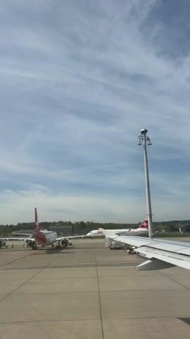 The video depicts an airport tarmac seen through an airplane window. In the foreground, part of the plane's wing is visible. In the middle distance, two Swiss airplanes are parked, attended to by ground crew. A tall light pole stands in the center of the view. The sky above is a light blue, with scattered wispy clouds. Beyond the tarmac, there is a treeline and some buildings.