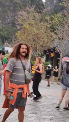 The video shows a crowded outdoor scene, possibly a tourist attraction. People of various ages and ethnicities are walking around. A man with curly hair and a mustache is prominent in the first shot, wearing a striped shirt and carrying a bag. Other people are dressed in casual clothing. There's a coffee shop visible in the background and trees lining the area. The ground is paved with bricks. In subsequent shots, more people are seen strolling around, some wearing traditional clothing. Red heart-shaped balloons are visible in one of the shots. The general atmosphere seems relaxed and bustling with activity.