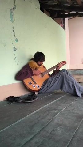 A man with dark hair and a beard is sitting on a wooden floor leaning against a light green wall. He is wearing a dark hoodie and a yellow scarf. He is playing a wooden acoustic guitar. There are two pairs of shoes on the floor to his left. The wall has a large crack running down its center. The ceiling above is made of dark wood.