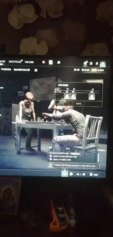 The video shows a screen displaying a video game. In the foreground, a person is sitting at a table, leaning over it. Another person is standing on the opposite side of the table, looking down at the person sitting. A menu is visible on the right side of the screen, displaying what appears to be weapon options or upgrades. Below the menu, there are text prompts in Russian, indicating game instructions or actions. The overall environment appears to be indoors, with dimly lit walls and some objects visible in the background.